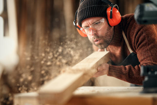 Adult male woodworker in protective goggles and headphones blowing sawdust from wooden detail while working in carpentry workshop